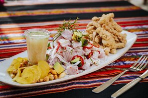 Een bord ceviche met avocado, rode peper en ui, een glas drinken en gebakken plantanen op een kleurrijke tafelkleed.