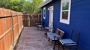 A patio with brick flooring, a wooden fence, and a blue building with a window and door. - Small Cozy Quite House (Wylie)