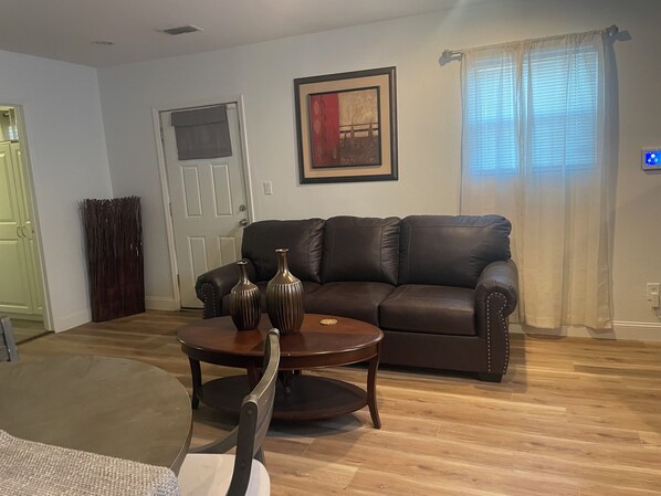 A living room with a brown leather sofa, a round wooden coffee table, and two metallic vases. There is a door with a window, a framed picture on the wall, and a curtain covering a window. - Small Cozy Quite House (Wylie)