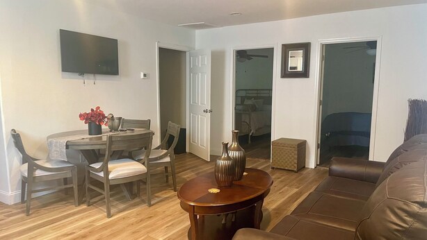 A living room with a brown leather couch, a wooden dining table with chairs, a coffee table, and a flat-screen TV mounted on the wall. - Small Cozy Quite House (Wylie)