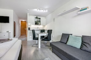 Une pièce moderne et bien éclairée, avec un lit, un canapé gris, un coin repas avec des chaises et une cuisine à l’arrière-plan.