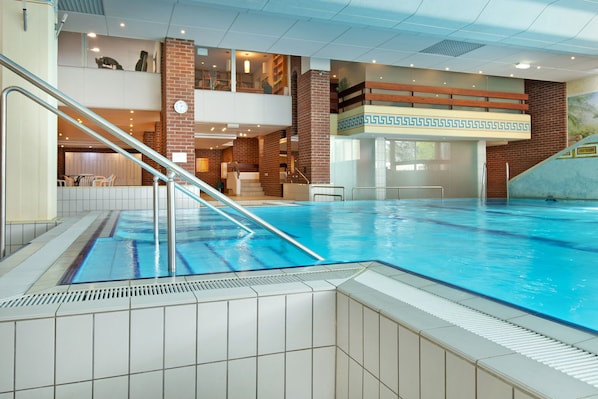 Une piscine intérieure avec un escalier, un balcon au deuxième étage et un mur en briques orné d’un motif décoratif.