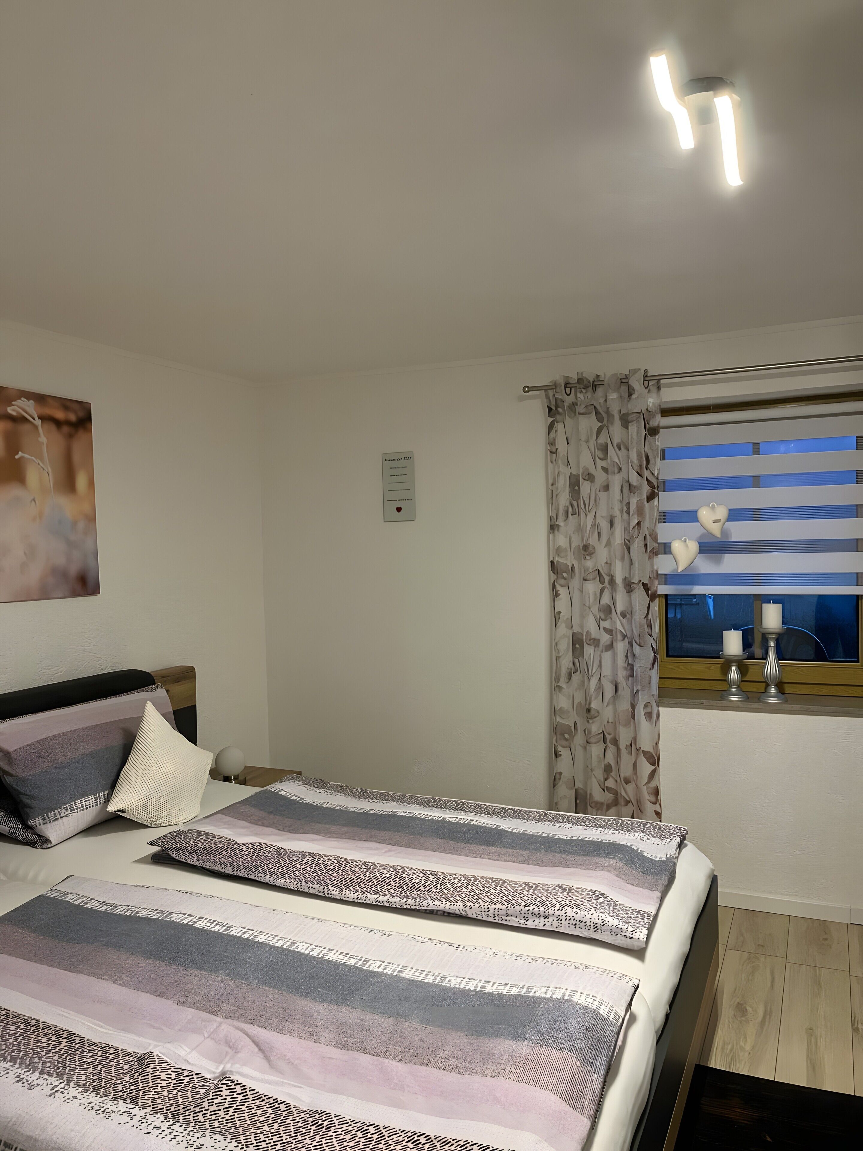 A bedroom with a bed, a window with curtains, a wall-mounted light fixture, and a framed picture.