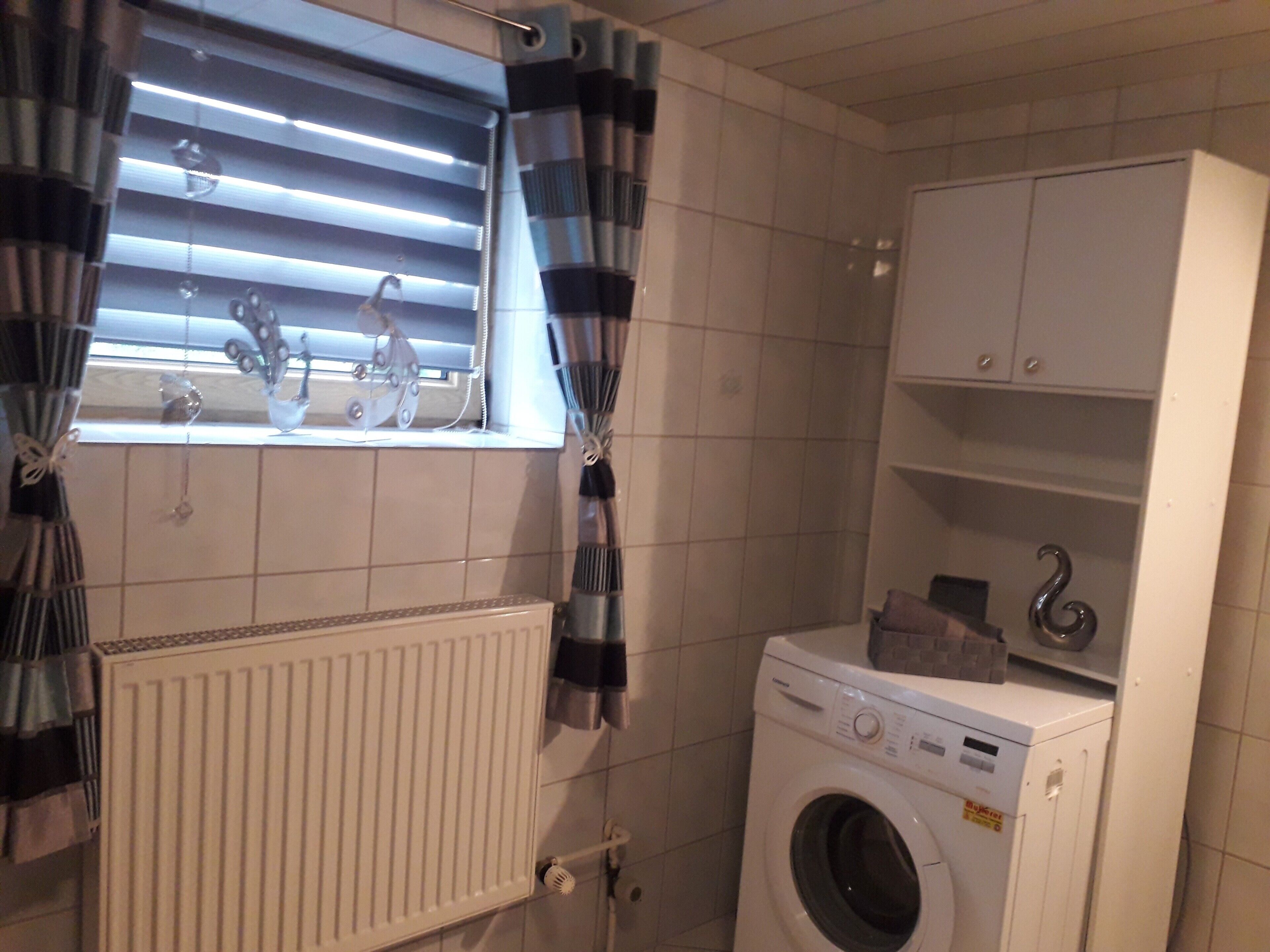 Une buanderie équipée d’un lave-linge, d’un radiateur et d’une fenêtre avec des stores.