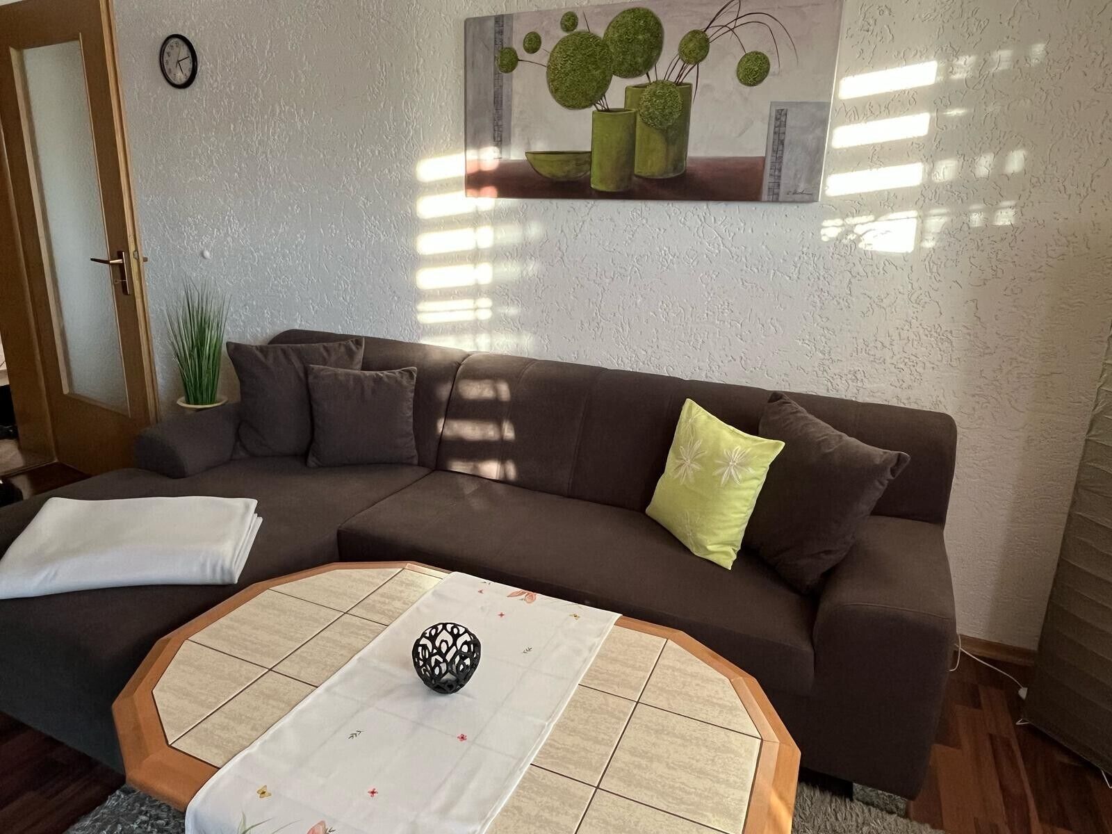 Uma sala de estar com um sofá marrom, uma mesa branca com um vaso estampado e um quadro na parede.