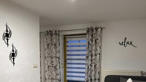 Een slaapkamer met een bed, een nachtkastje, een raam met gordijnen, een muur met een ‘relax’ bord en een plafondlamp.