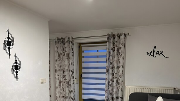 A bedroom with a bed, a bedside table, a window with curtains, a wall with a 'relax' sign, and a ceiling light fixture. - Holiday Apartment "Ferienwohnung Köppl" with Terrace (LAM)