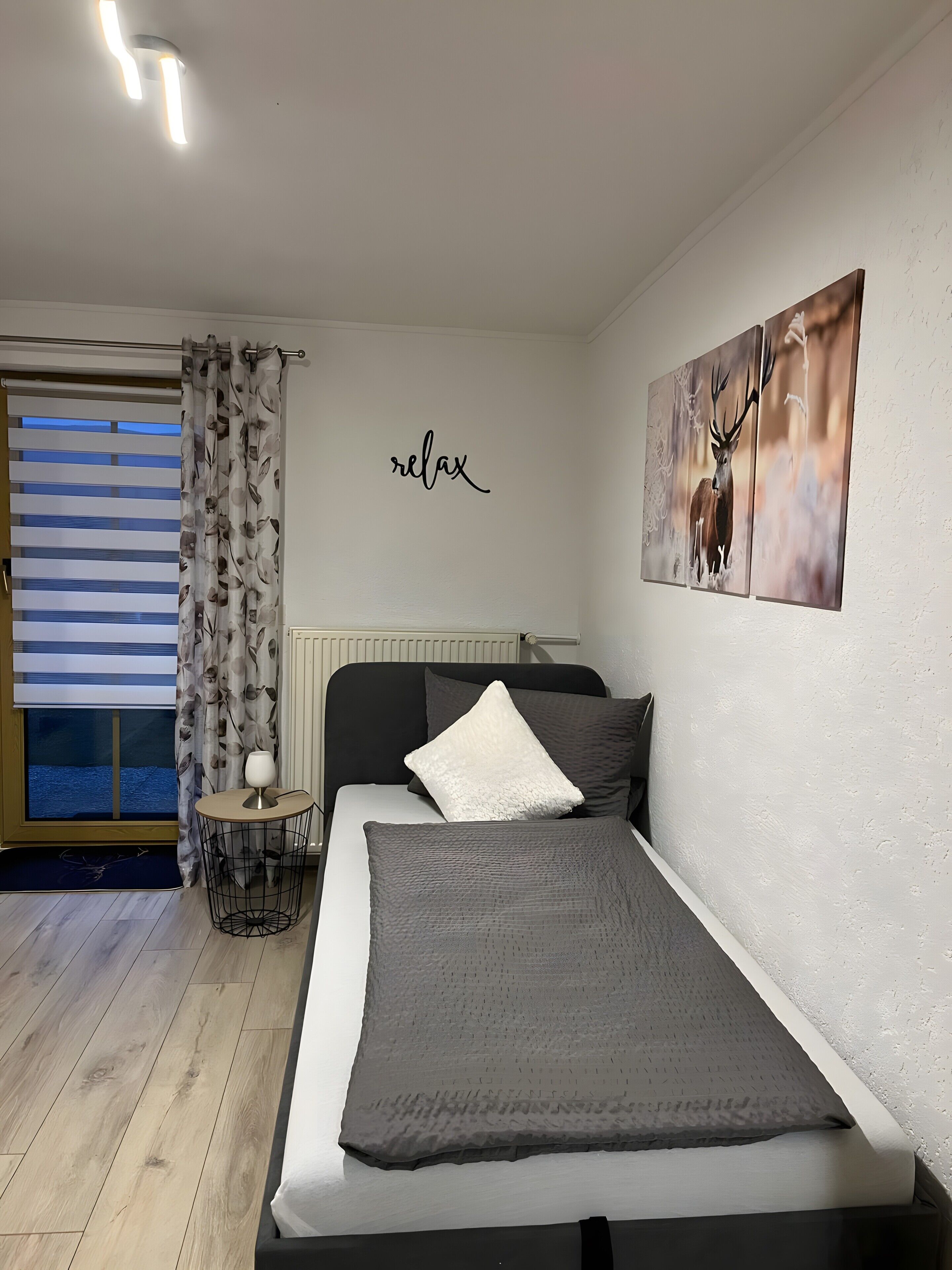 A bedroom with a bed, a bedside table with a lamp, a window with blinds, a wall with a 'relax' sign, and a deer painting.