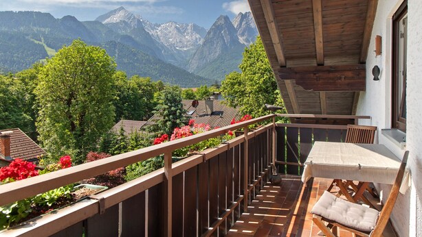 A balcony with a wooden railing, a table with a cushion, and a view of mountains. - Holiday Apartment "Anette" with Mountain View, Balcony & Wi-Fi (Garmisch-Partenkirchen)