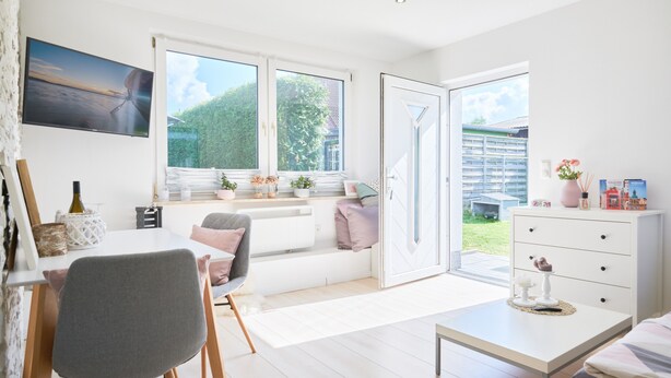 Une pièce lumineuse avec une grande fenêtre, un coin repas, un canapé et une commode blanche.