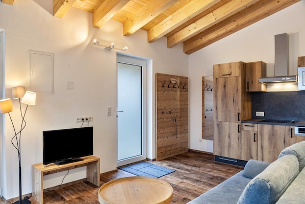 A modern living room with a television on a wooden stand, a sofa, a dining table, and a kitchen area with wooden cabinets and a microwave.