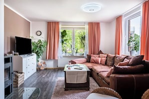 En stue med sofa, fjernsyn, ventilator, et vindue med gardiner og et ur på væggen.