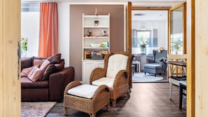 Een woonkamer met een bruine leren bank, een rieten fauteuil met kussen, een witte boekenkast, en een eethoek met tafel en stoelen.