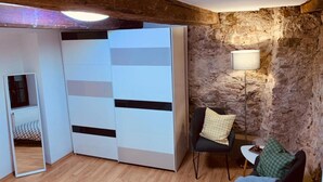 A room with a wooden ceiling, a stone wall, a white wardrobe, a black chair, a small table, and a lamp.