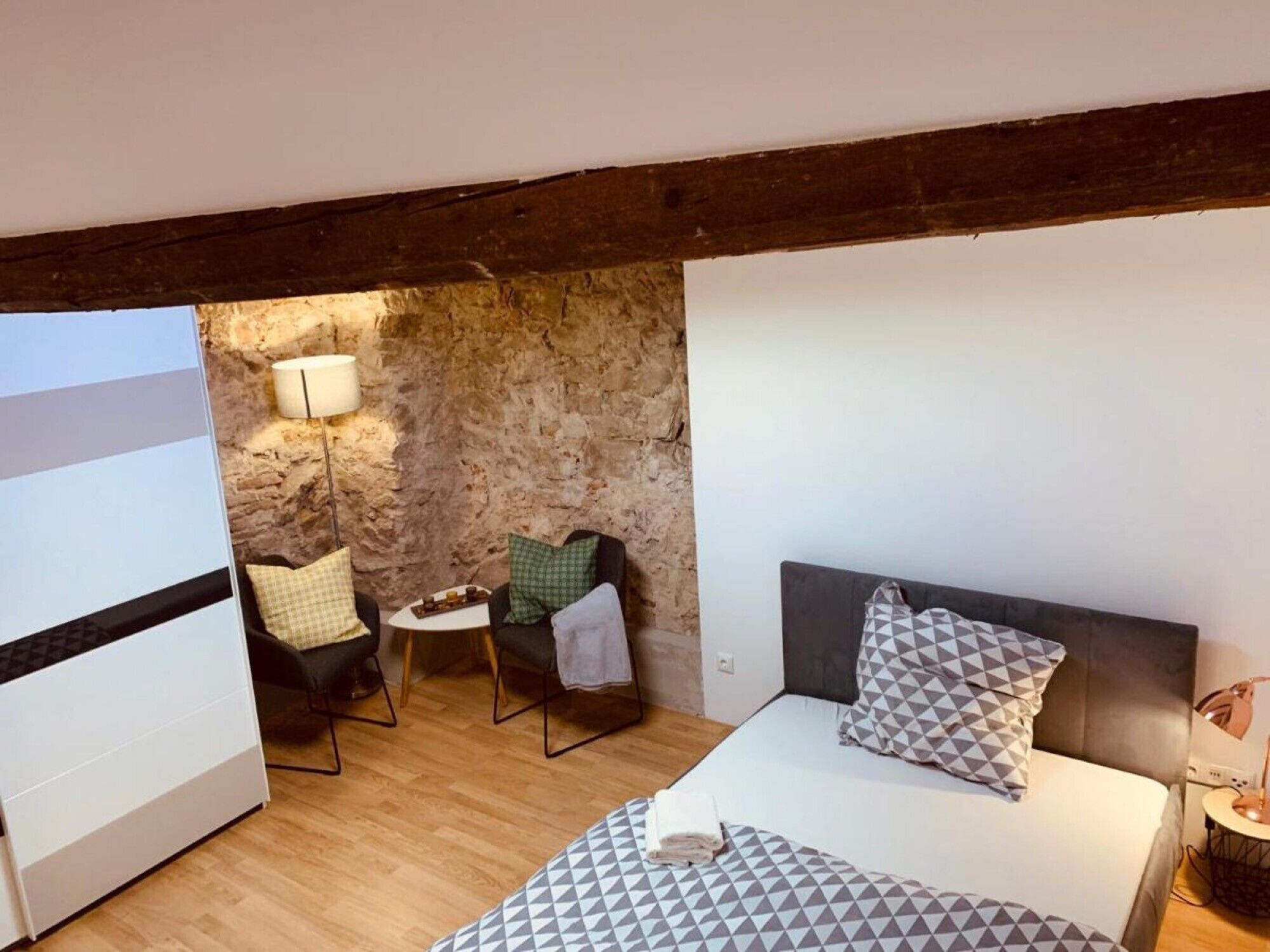 Ein Schlafzimmer mit Bett, Stuhl, kleinem Tisch, Lampe und einer Steinwand.