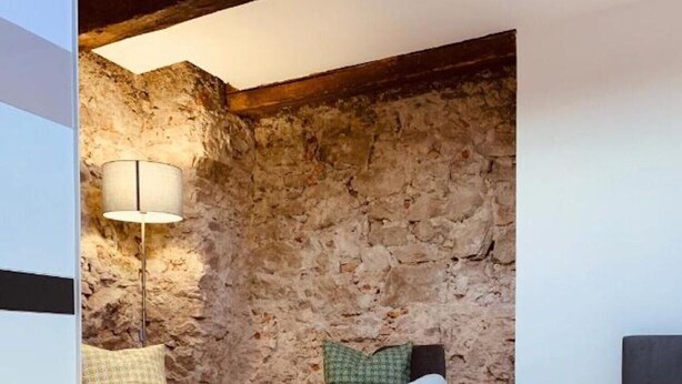 A room with a stone wall, a bed with a geometric patterned bedspread, two chairs with cushions, and a floor lamp.