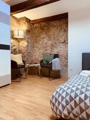 Ein Zimmer mit Steinsandsteinwand, einem Bett mit geometrischem Muster auf der Bettdecke, zwei Sesseln mit Kissen und einer Stehlampe.