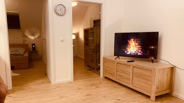 Une pièce avec un meuble TV en bois, un téléviseur à écran plat diffusant une image de cheminée, une horloge au mur et une table en verre.