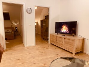 Une pièce avec un meuble TV en bois, un téléviseur à écran plat diffusant une image de cheminée, une horloge au mur et une table en verre.