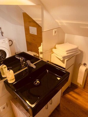 Une petite salle de bain avec un lavabo noir, un miroir, un porte-serviettes et un lave-linge.