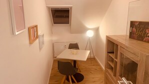 Une petite pièce étroite, avec une table blanche, deux chaises, un radiateur, un luminaire au plafond et un meuble en bois doté d’une porte en verre.