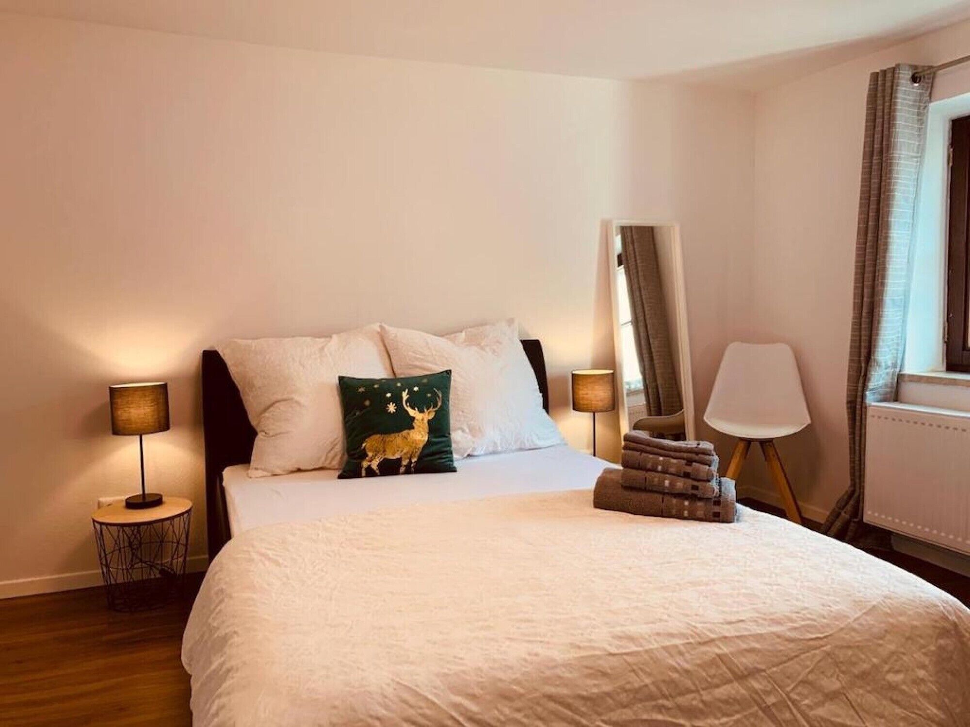 A hotel room with a bed, two bedside lamps, a chair, a mirror, and a window with curtains.