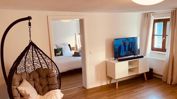 A room with a hanging chair, a bed with a nightstand, a TV on a white stand, and a window with curtains. - Holiday Apartment "Felix Living 2" with Wi-Fi (Passau)