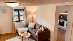 A living room with a sofa, a small table, a lamp, and a view of a kitchen through an open door. - Holiday Apartment "Felix Living 2" with Wi-Fi (Passau)