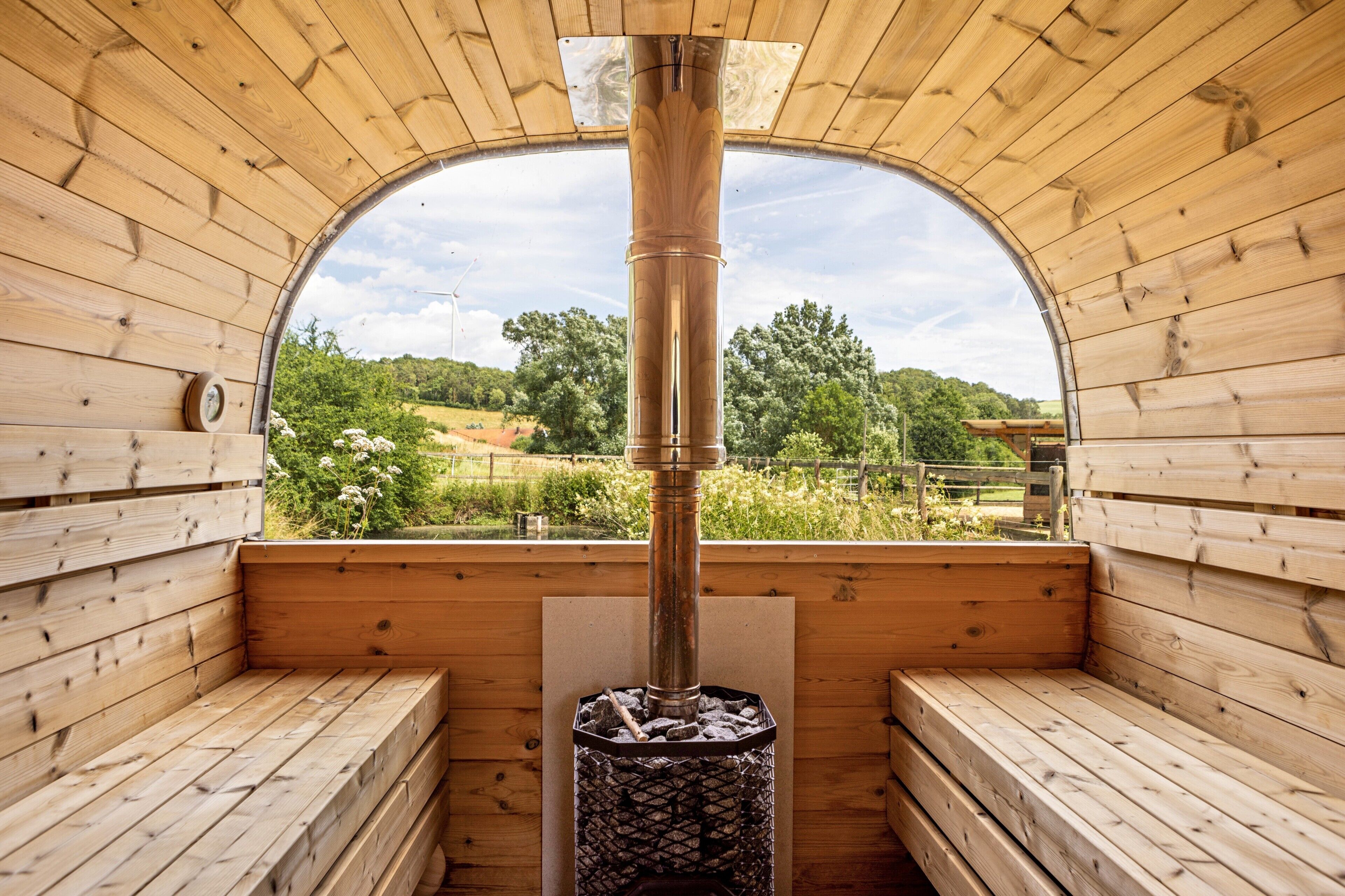 Una sauna con bancos de madera, una columna de calefacción central y vistas a la vegetación a través de un ventanal semicircular.