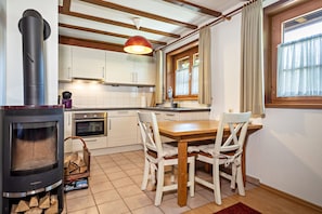 Une cuisine équipée d’un poêle à bois, de meubles blancs, d’une table à manger avec des chaises et d’une fenêtre avec des rideaux.