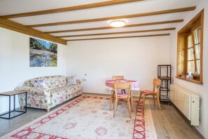 A room with a wooden ceiling, a sofa, a dining table with chairs, a radiator, and a window with a view of trees.