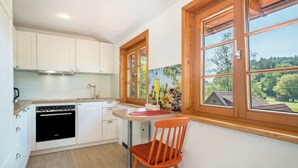 Une cuisine avec des meubles blancs, un four noir, une petite table recouverte d’un tapis rouge et une chaise en bois.