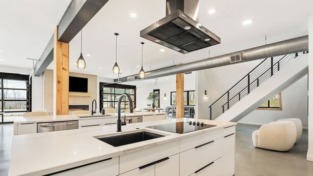 Una cocina moderna con una isla central, electrodomésticos de acero inoxidable, iluminación colgante y una escalera que sube a un nivel superior.