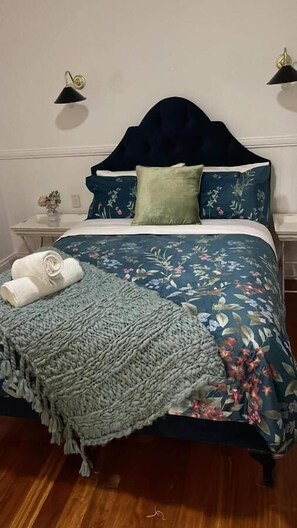 A neatly made bed with a floral bedspread, a dark headboard, and two wall-mounted lamps. - Emirau (Montville)