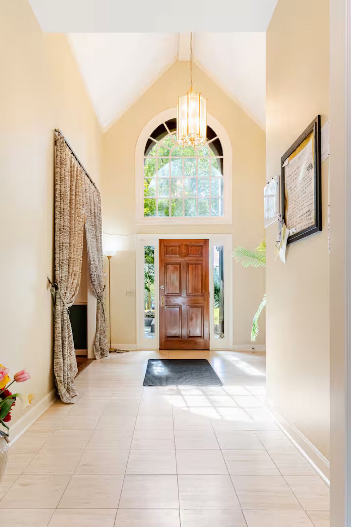 A hallway with a wooden door, a chandelier, and a view of greenery outside.