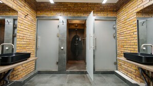 Baño público con paredes de ladrillo, dos lavamanos y una puerta que da a una zona de duchas.