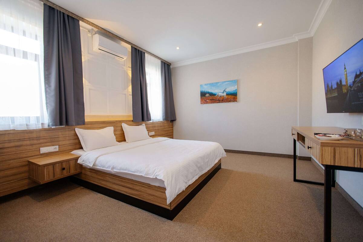 A modern hotel room with a large bed, wooden headboard, bedside tables, a desk, and a flat-screen TV.