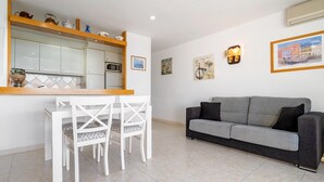 Ein modernes Wohnzimmer mit grauem Sofa, weißen Stühlen, einem Holzessttisch und einer Küchenzeile mit Schränken und einer Mikrowelle.