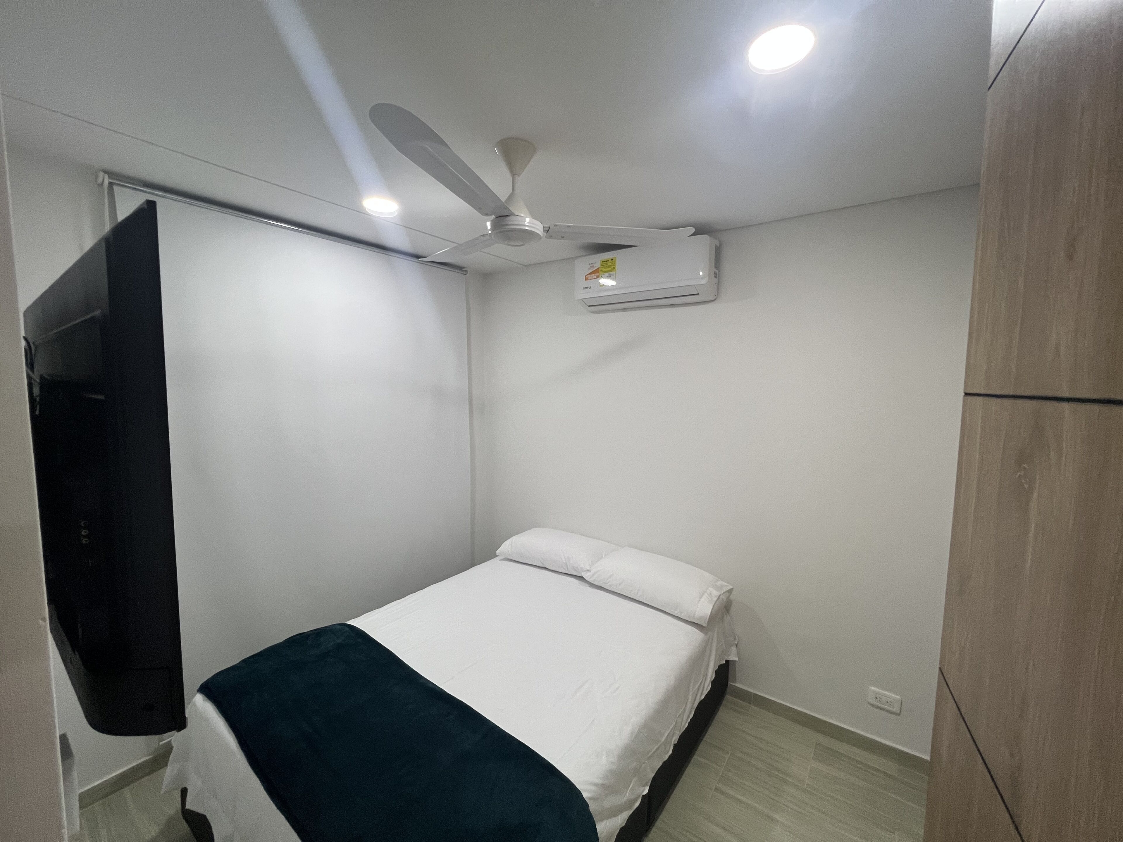 A bedroom with a bed, a ceiling fan, an air conditioning unit, and a TV mounted on the wall.