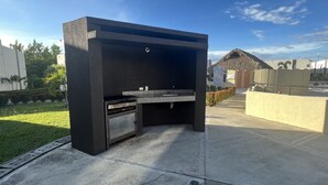 Een buitenkeuken met een ingebouwde oven en wasbak, omringd door een betonnen terras en een gazon.