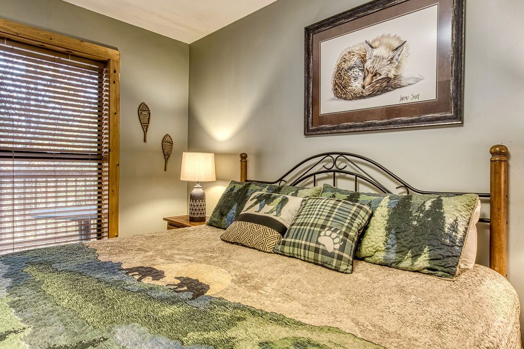 A bedroom with a bed, a nightstand, a lamp, a framed picture, and a window with blinds.