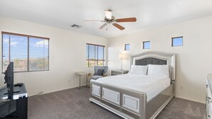 Ein Schlafzimmer mit einem Bett, einem Schreibtisch mit Computer, einem Deckenventilator und zwei Fenstern.