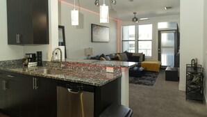 Cocina moderna con encimeras de granito, electrodomésticos de acero inoxidable y muebles de cocina negros. Hay un rincón de comedor con un sofá y una mesa de centro.