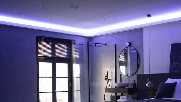 A modern bedroom with a large bed, a built-in bench, a bedside table with a mirror, and a window with blinds. - Ennéa - Jacuzzi & Luxury Suites (Perpignan)