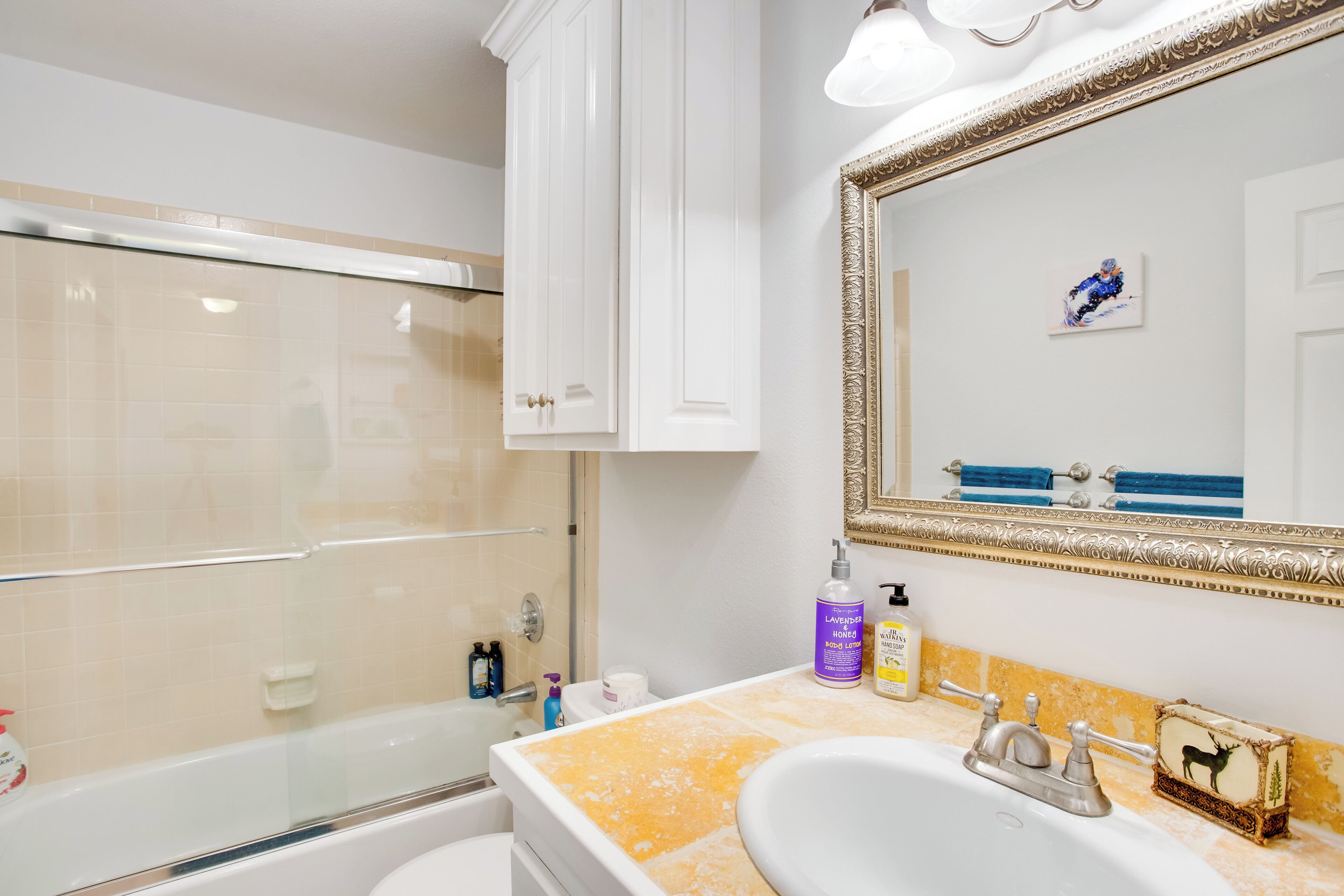 A bathroom with a glass shower enclosure, a sink with a gold and white tiled countertop, and a large mirror with an ornate frame.