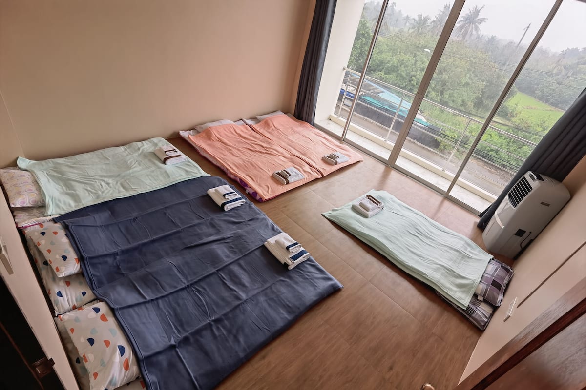 A room with three beds, each with a different colored bedspread and pillows, and a large window with a view of greenery and a building outside.