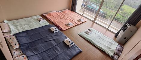 A room with three beds, each with a different colored bedspread and pillows, and a large window with a view of greenery and a building outside.