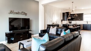 A modern living room with a large flat-screen TV, a black leather sofa with patterned cushions, and a dining area with a kitchen in the background.