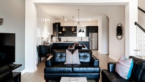 A modern living room with a black leather sofa, a flat-screen TV, and a dining area with a kitchen in the background.