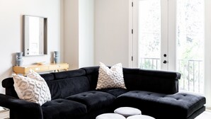 Un canapé d’angle noir agrémenté de deux coussins décoratifs à motifs, une petite table d’appoint en bois et un miroir placé au-dessus.